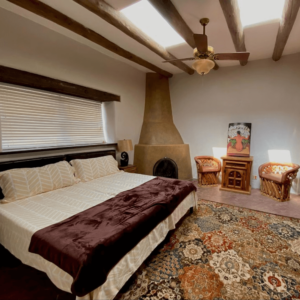 Spacious bedroom with a king-sized bed dressed in soft, neutral linens and a cozy brown throw, featuring an adobe-style fireplace, rustic wooden beams, skylights, and two handcrafted chairs with a small decorative table for a Southwestern charm.