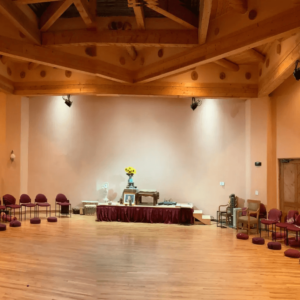 Spacious meditation hall with warm wooden flooring and a serene altar adorned with sunflowers, surrounded by neatly arranged chairs and cushions, under a rustic wooden ceiling.