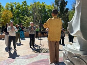 A group of individuals practicing mindfulness and meditation under the guidance of Master Mingtong Gu in a golden robe, gathered outdoors near a serene statue on a sunny day with lush greenery in the background.