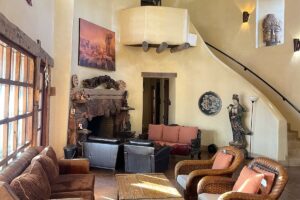 Cozy living room featuring rustic decor with a stone fireplace, wicker chairs, a Buddha wall sculpture, and a serene statue of a female figure, illuminated by warm natural light.