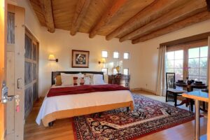 Cozy bedroom with a Southwestern design, featuring a wooden beam ceiling, a large bed with vibrant bedding, a patterned rug, and natural light streaming through windows and French doors.