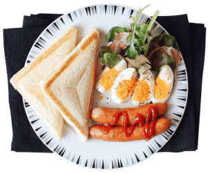 Plate of breakfast food featuring toasted sandwich bread, boiled eggs, fresh greens, and sausages topped with ketchup, presented on a white plate with black patterned edges.