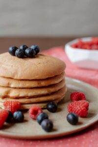 Stack of fluffy pancakes topped with fresh blueberries and surrounded by raspberries, served on a rustic plate with a blurred background