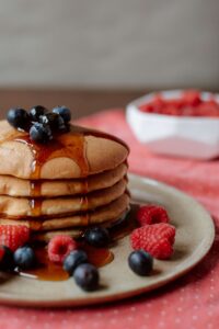 A stack of fluffy pancakes topped with fresh blueberries, drizzled with maple syrup, and surrounded by vibrant raspberries, served on a rustic beige plate with a bowl of additional raspberries in the background