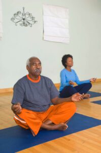 Two individuals sitting cross-legged on yoga mats, meditating in a serene indoor space with a light-colored wall and natural wood flooring.
