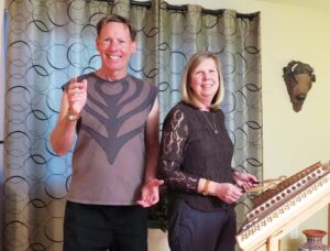 Bill and Laurie posing indoors, with Bill smiling and gesturing while Laurie stands beside a hammered dulcimer, ready to play, set against a patterned curtain backdrop.