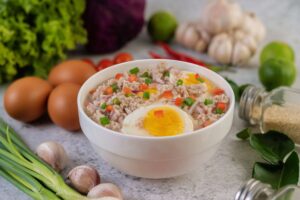 Warm bowl of savory rice porridge topped with minced pork, hard-boiled eggs, diced carrots, and green onions, surrounded by fresh ingredients like garlic, eggs, and vegetables.