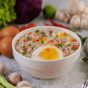 Warm bowl of savory rice porridge topped with minced pork, hard-boiled eggs, diced carrots, and green onions, surrounded by fresh ingredients like garlic, eggs, and vegetables.