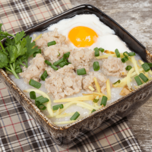 Traditional rice porridge topped with minced pork, a sunny-side-up egg, ginger strips, fresh cilantro, and green onions, served in a rustic bowl on a plaid cloth.