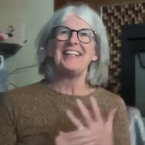 Jani smiling brightly, wearing glasses and a textured brown top, gesturing warmly with her hand on her chest, set in a cozy indoor environment with subtle decorative elements in the background.