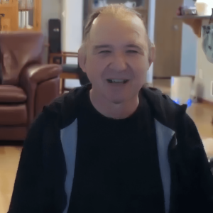 John Maynes smiling warmly, seated indoors in a cozy living room with soft lighting and comfortable furniture in the background.