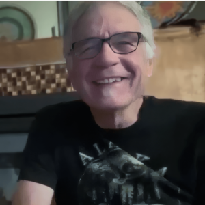 John Moore smiling warmly while wearing glasses and a black shirt, set against a cozy indoor background.