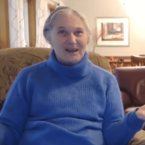 Judy sitting in a cozy living room, wearing a bright blue turtleneck sweater, speaking warmly with a gesture, with soft natural light and framed artwork in the background.