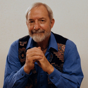 Len Schreiner seated with a calm and thoughtful demeanor, wearing a blue shirt and patterned vest, radiating wisdom and warmth.