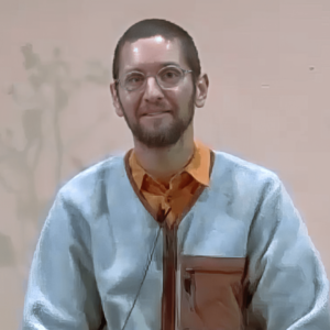 Luke wearing glasses and a light blue sweater over an orange shirt, sitting in a calm and neutral setting with a soft background, offering a warm and thoughtful expression.