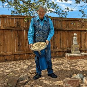 Rick Jan smiling while playing a blue drum in an outdoor setting with a wooden fence and decorative statue in the background, showcasing joy and musical expression.