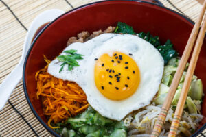 Delicious bibimbap dish featuring a fried egg topped with black sesame seeds, shredded carrots, spinach, cucumber, bean sprouts, and rice, served in a red bowl with chopsticks.