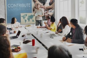 Team collaborating in a modern conference room with a speaker presenting ideas under a 'Contribute' banner, fostering global engagement and teamwork with a world map display in the background.