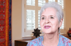 Bianca shares her Wisdom Healing Qigong journey, her face radiating calmness and wisdom. She is seated in a warm, inviting space adorned with colorful wall hangings and natural light streaming through nearby windows.