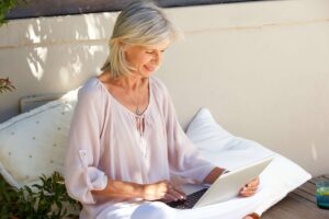 Smiling mature woman with silver hair working on a laptop while sitting outdoors on a cozy cushion, enjoying natural light and a relaxed setting.
