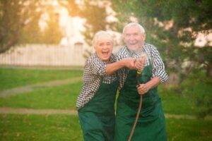 Elderly couple laughing joyfully while playfully spraying water with a garden hose, wearing matching green aprons and plaid shirts.