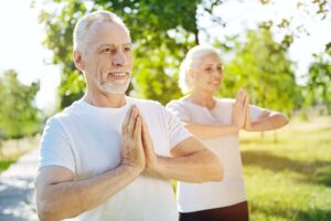 Smiling senior couple practicing yoga or meditation outdoors in a sunny park, standing with hands in a prayer position.