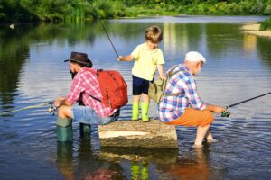 Three generations fishing together by a calm lake: a grandfather, father, and young boy with fishing rods and backpacks, enjoying a sunny day outdoors.
