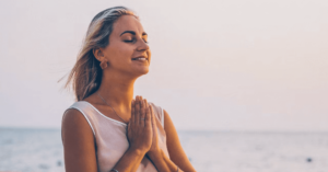 Smiling woman with hands in prayer position by the ocean, embracing mindfulness and gratitude at sunrise.