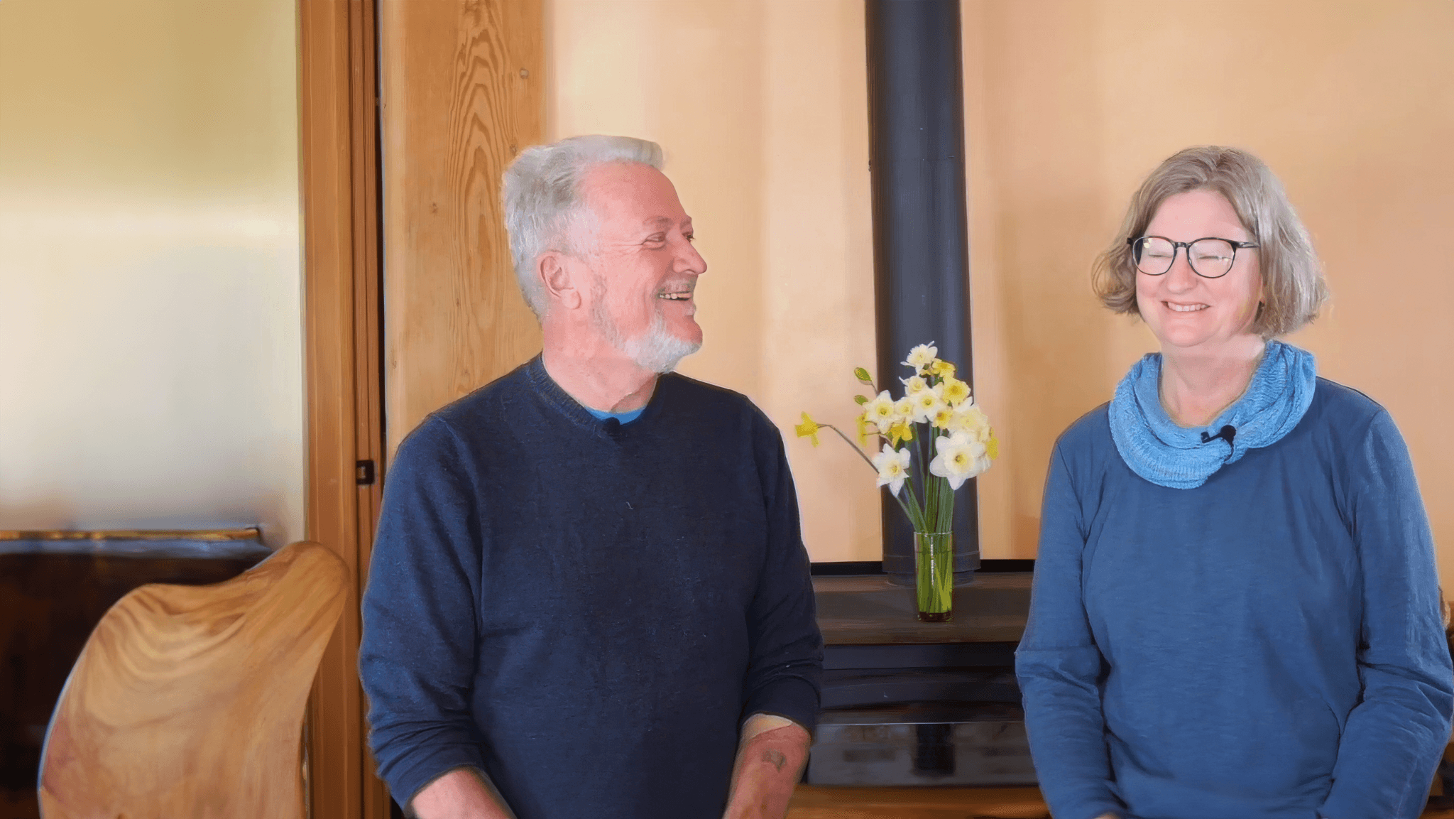 Smiling man and woman sitting indoors in a cozy setting, with a vase of yellow flowers on a table and a wood-paneled background.
