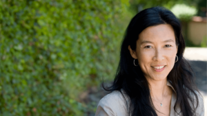 Portrait of a smiling woman with long black hair, wearing hoop earrings and a necklace, standing outdoors with a lush green background.