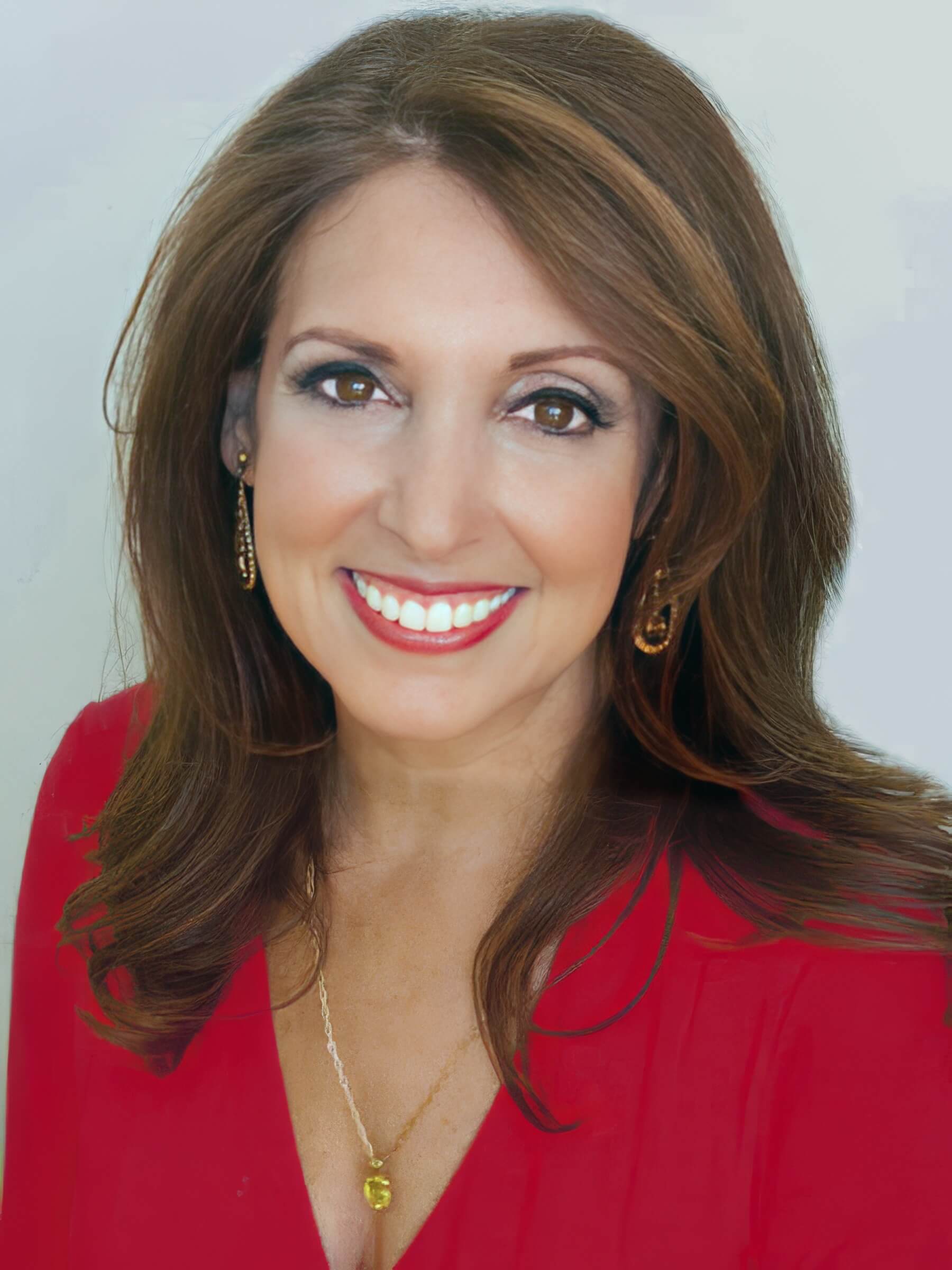 Portrait of Marci Shimoff, smiling brightly, wearing a red top with gold earrings and a matching necklace, set against a light background.