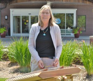 Laurie Nelson stands outside, smiling as she plays a hammered dulcimer. She is wearing a light cardigan and a turquoise necklace, adding a serene touch to the natural greenery and flowers surrounding her.