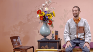 Luke shares his Wisdom Healing Qigong experience, seated comfortably with a relaxed smile beside a vibrant floral arrangement, embodying a calm and welcoming presence.