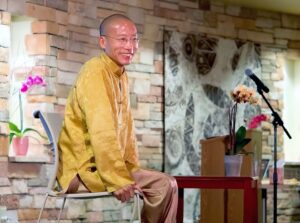 Master Mingtong Gu, dressed in a vibrant golden robe, sits gracefully on a chair with a warm, gentle smile. The backdrop of textured stone and soft, earthy decor creates a calming atmosphere. A potted orchid and a microphone are positioned nearby, adding to the serene and inviting setting for his Wisdom Healing Qigong teaching.