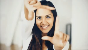 A woman smiling brightly as she frames her face with her fingers, creating a square shape with her hands, symbolizing focus and clarity. Her expression radiates positivity, suggesting an open and mindful perspective, aligning with themes of self-awareness and intentionality in energy medicine.