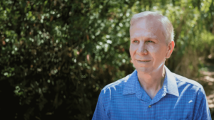 Tom stands outdoors, softly lit by natural sunlight, surrounded by lush greenery that creates a calm and reflective atmosphere. His serene expression and relaxed posture convey a sense of peace and well-being, perfect for a Wisdom Healing Qigong testimonial.