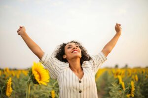 Joyful woman with curly hair stands in a field of sunflowers, raising her arms triumphantly toward the sky, radiating a sense of freedom and vitality under a bright, clear sky.