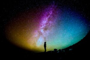 A solitary figure stands silhouetted against a breathtakingly vibrant Milky Way galaxy, painted in a spectrum of blues, pinks, and yellows. This celestial scene inspires a sense of awe and is emblematic of the cosmic energy and vastness often contemplated in Wisdom Healing Qigong practice.