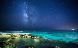 A serene night sky, speckled with countless stars, featuring the Milky Way stretching above a tranquil, glowing turquoise sea. The foreground shows rugged rocky outcrops at the water's edge, enhancing the peaceful yet awe-inspiring ambiance of the scene, reflecting the meditative and cosmic connection emphasized in Wisdom Healing Qigong practice.