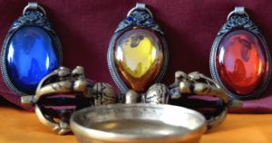 Three vibrant gemstone pendants in detailed silver settings—blue, yellow, and red—displayed alongside an ornate metal bowl, against a deep burgundy background, evoking a sense of spiritual energy and mysticism.