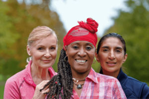 Three senior women from diverse backgrounds smiling together, highlighting the unity and community of Wisdom Healing Qigong practices. The Caucasian, African, and Hispanic women are outdoors, with a backdrop of lush greenery, reflecting the serene environment that supports their wellness journey.