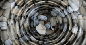 A mesmerizing swirl of smoothed river stones in varying shades of gray, brown, and white, forming a circular pattern that symbolizes the continuous flow of source energy associated with Hun Yuan Chi. This natural mandala evokes the harmonious principles of Wisdom Healing Qigong and sound healing, reflecting balance and tranquility.