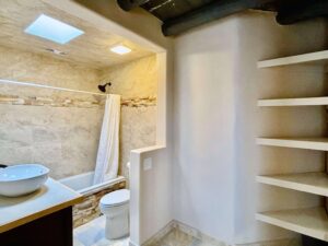 Modern bathroom in a loft room at The Chi Center, designed to complement Wisdom Healing Qigong practice with a serene and clean atmosphere. The bathroom features beige stone tiles, a shower with a white curtain, a vessel sink on a wooden countertop, and built-in white shelves. Skylights provide natural light, enhancing the calm and inviting space.