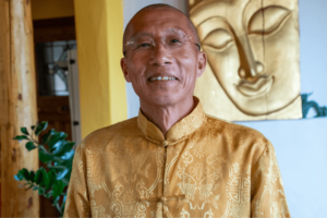Portrait of Master Mingtong Gu in a golden yellow robe adorned with detailed patterns, smiling gently, with glasses, in front of a large golden Buddha face. The warm indoor lighting enhances the serene and cultural ambiance of the setting.