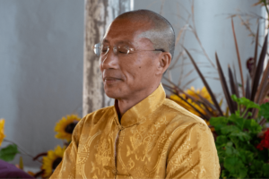 A serene portrait of Master Mingtong Gu meditating, eyes closed, dressed in a traditional golden yellow robe with intricate patterns. The backdrop is softly blurred with lush greenery and colorful flowers, contributing to the peaceful and spiritual ambiance.