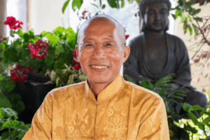 Portrait of a joyful Master Mingtong Gu in glasses, dressed in a traditional golden yellow robe, smiling warmly in a lush indoor garden setting with a Buddha statue in the background.