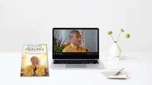 A minimalist workspace setting featuring a laptop playing a video of Master Mingtong Gu in a yellow robe, with the E-Book 'Sound Healing Technologies' and a simple white vase with green flowers. The clean and serene environment highlights the theme of sound healing and mindfulness.