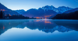 A tranquil pre-dawn scene captures the serene blue waters of a lake perfectly reflecting the surrounding snow-capped mountains and the warm glow of village lights. The peaceful and reflective atmosphere provides an ideal setting for Wisdom Healing Qigong practice, nurturing a state of pure consciousness amidst nature's beauty.