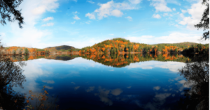 Panoramic view of a serene lake surrounded by a forest in full autumn splendor, reflecting a clear blue sky and fluffy clouds. The vibrant orange and red foliage on the hills around the lake offers a peaceful and revitalizing setting for practicing Wisdom Healing Qigong, promoting a deep connection with nature and nurturing pure consciousness.