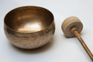 Traditional Tibetan singing bowl and wooden mallet on a white background, representing tools used in sound healing and Wisdom Healing Qigong practices. The golden bowl, marked with patina, is associated with meditation and the therapeutic power of sound, emphasizing tranquility and healing.